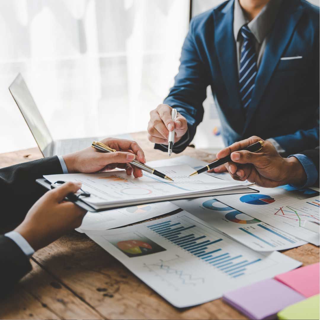 Three men are seated around a rustic wooden table, each holding a pen and attentively reviewing financial data presented on a clipboard held by one of them.