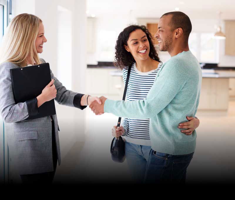 A female realtor holding a clipboard is shaking hands with a smiling couple in an empty well lit house.
