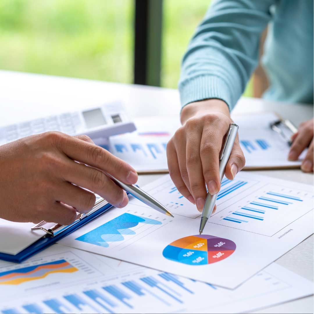 A man and a woman are seated at a desk across from each other, using pens to point at financial data printed on sheets of paper in a well-lit office space.